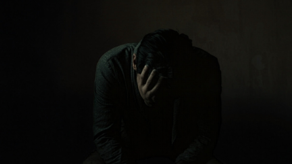How Much Adderall Is Too Much hero image of a man holding his head while sitting in the dark.